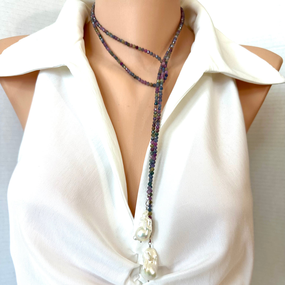 A close-up of a beaded lariat-style necklace with large white pearl-like beads, worn around the neck of a model wearing a white sleeveless blouse.