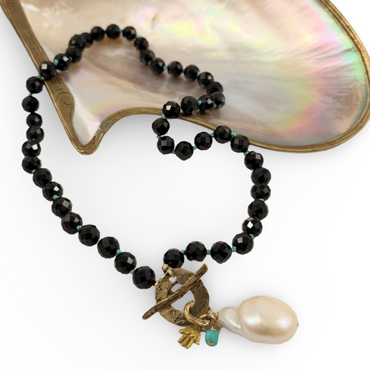 A beaded necklace with faceted black beads, a small turquoise accent, and a toggle clasp featuring a baroque pearl and gold-toned details, displayed on a white background with a mother-of-pearl shell prop.