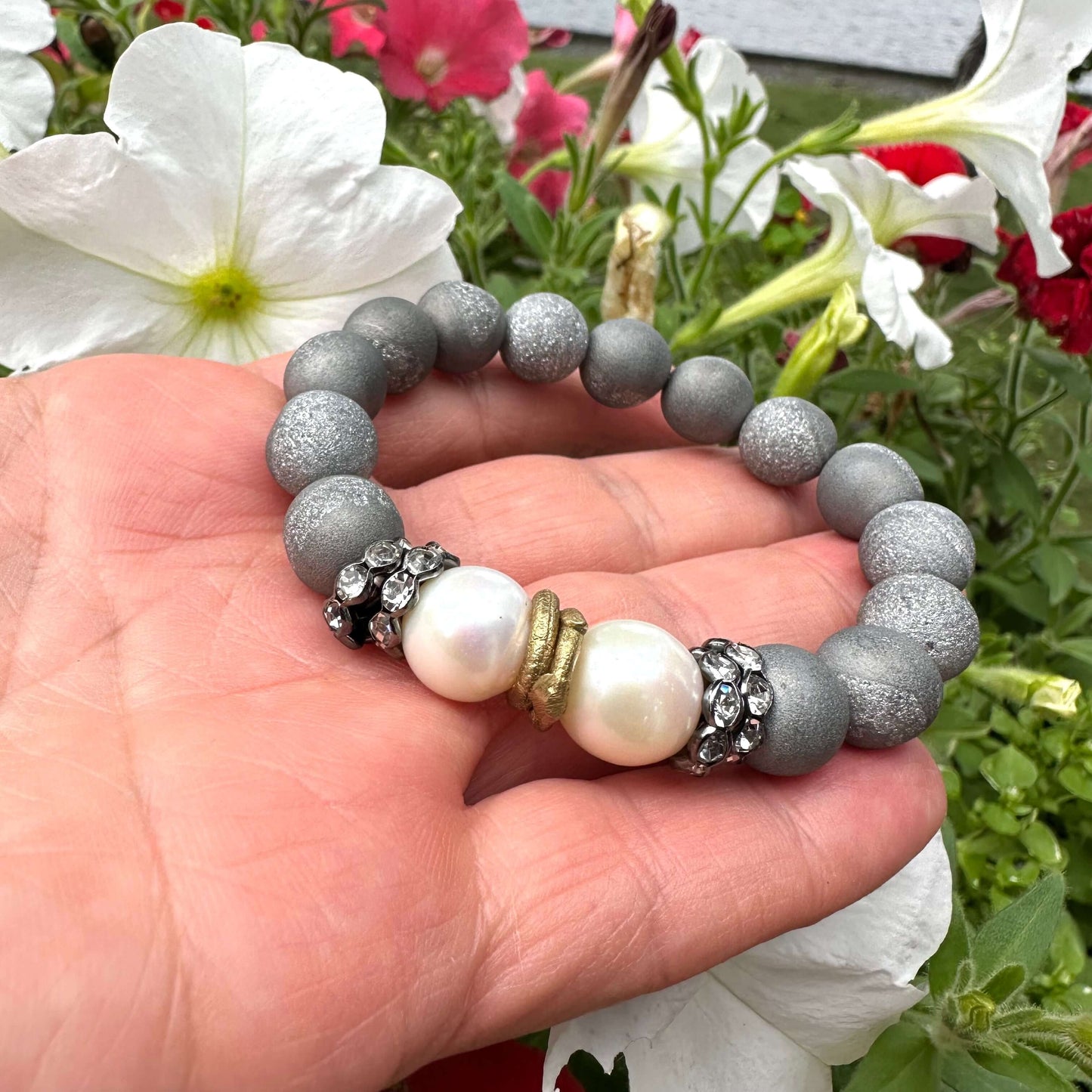 Close-up of grey druzy agate and pearl bracelet held outdoors near foliage