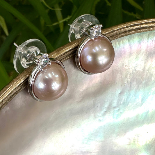 Handcrafted pink mabe pearl stud earrings resting on white shell with soft greenery, showing luminous blush hue and silver setting.