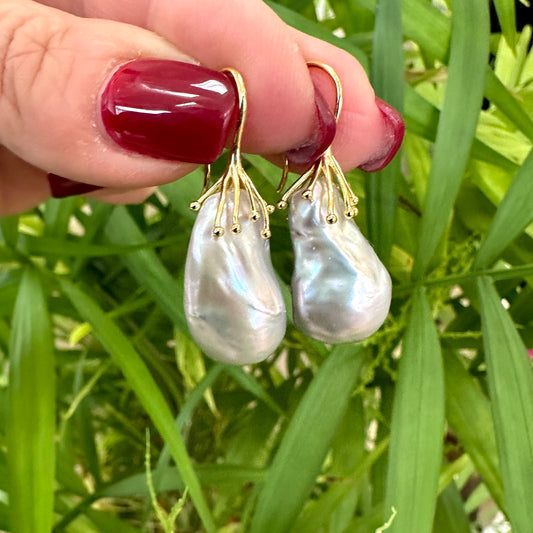 Gold vermeil silver grey baroque pearl drop earrings held in hand against green foliage, highlighting luminous silver-grey hue.