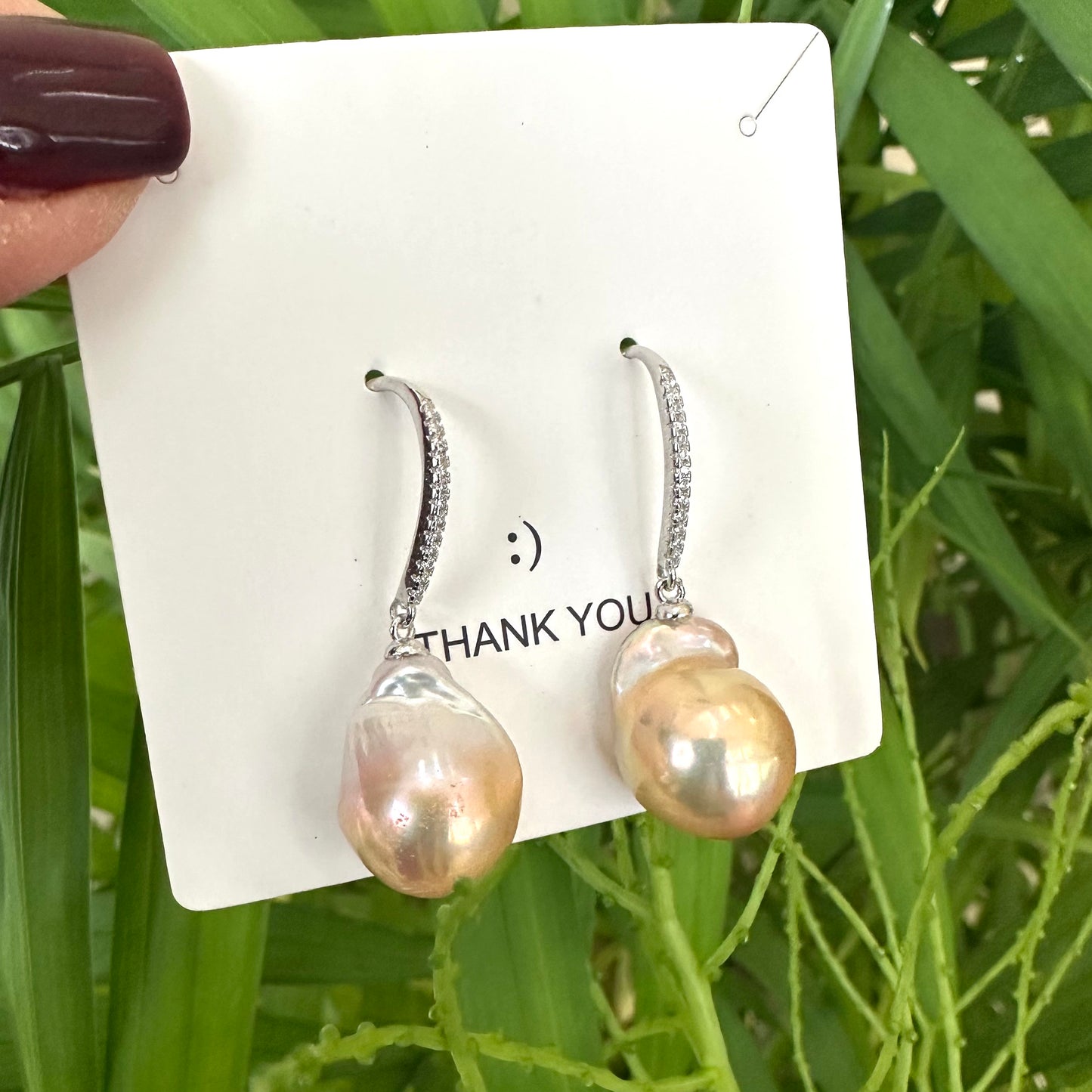 Golden pink baroque pearl dangle earrings displayed against green leaf backdrop, designed and handmade in Canada.