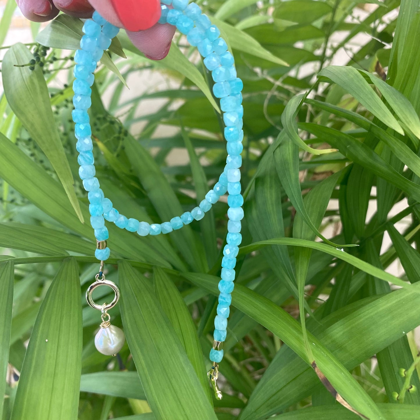 Blue amazonite baroque pearl necklace showcasing aqua-blue gemstones and charm design held amongst green leaves.