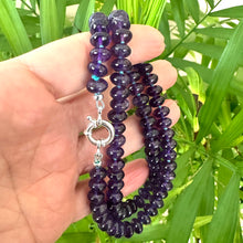 Load image into Gallery viewer, Amethyst beaded necklace held in hand for size, against green leaf background