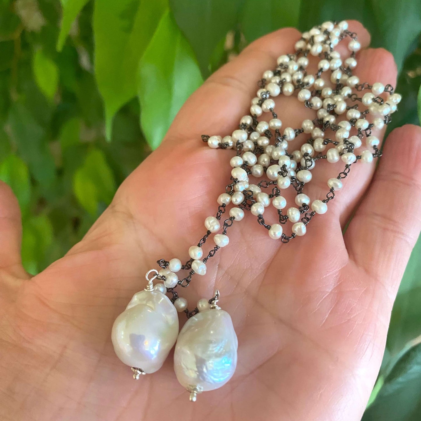 Pearl lariat rosary chain necklace held in hand against green foliage showing pearl luster