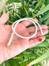 Load image into Gallery viewer, Handcrafted White Pearl Necklace featuring red heart charm and gold clasp held near green leaf background.