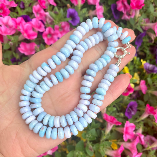 opal candy necklace in pinkish blue tones featuring handcrafted rondelle beads on a hand against a backdrop of colorful flowers.