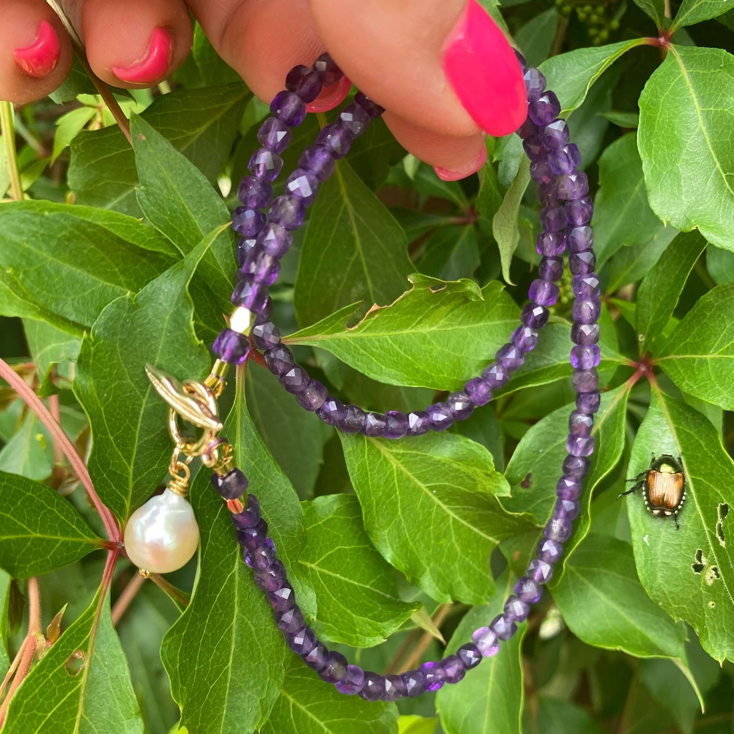 Amethyst toggle necklace with baroque pearl charm displayed against green foliage.