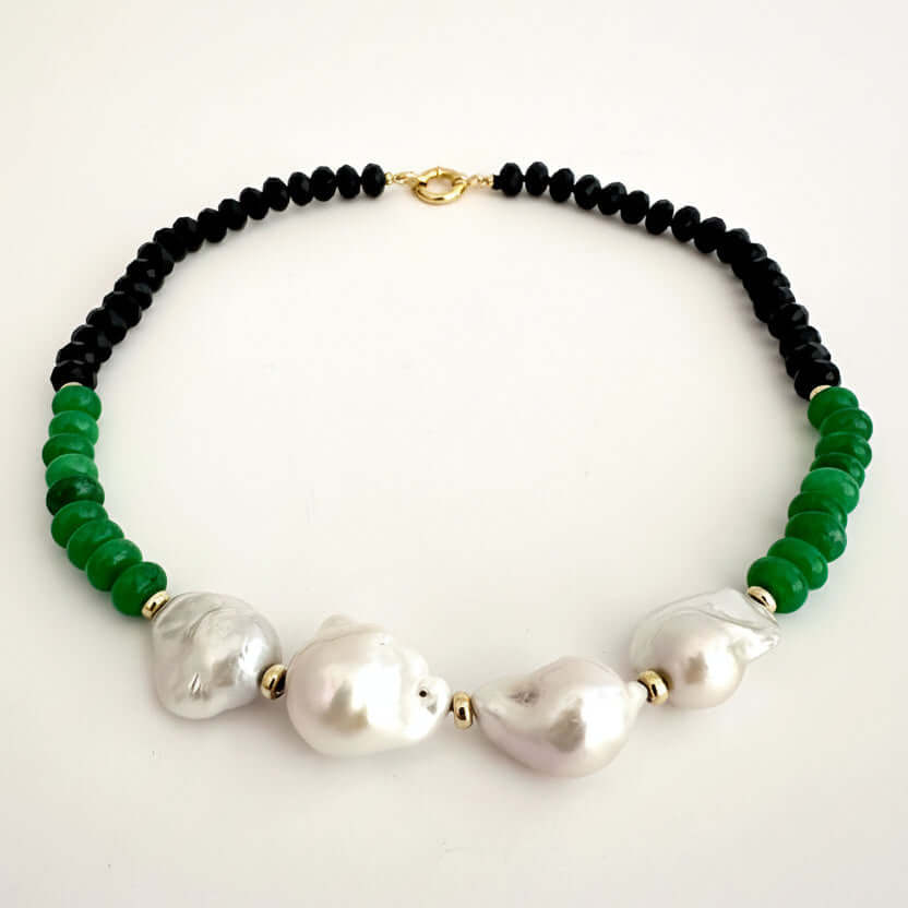 A beaded necklace featuring black round beads, vibrant green faceted beads, and three large white baroque pearls, accented with small gold spacers and a gold clasp, displayed on a clean white background.