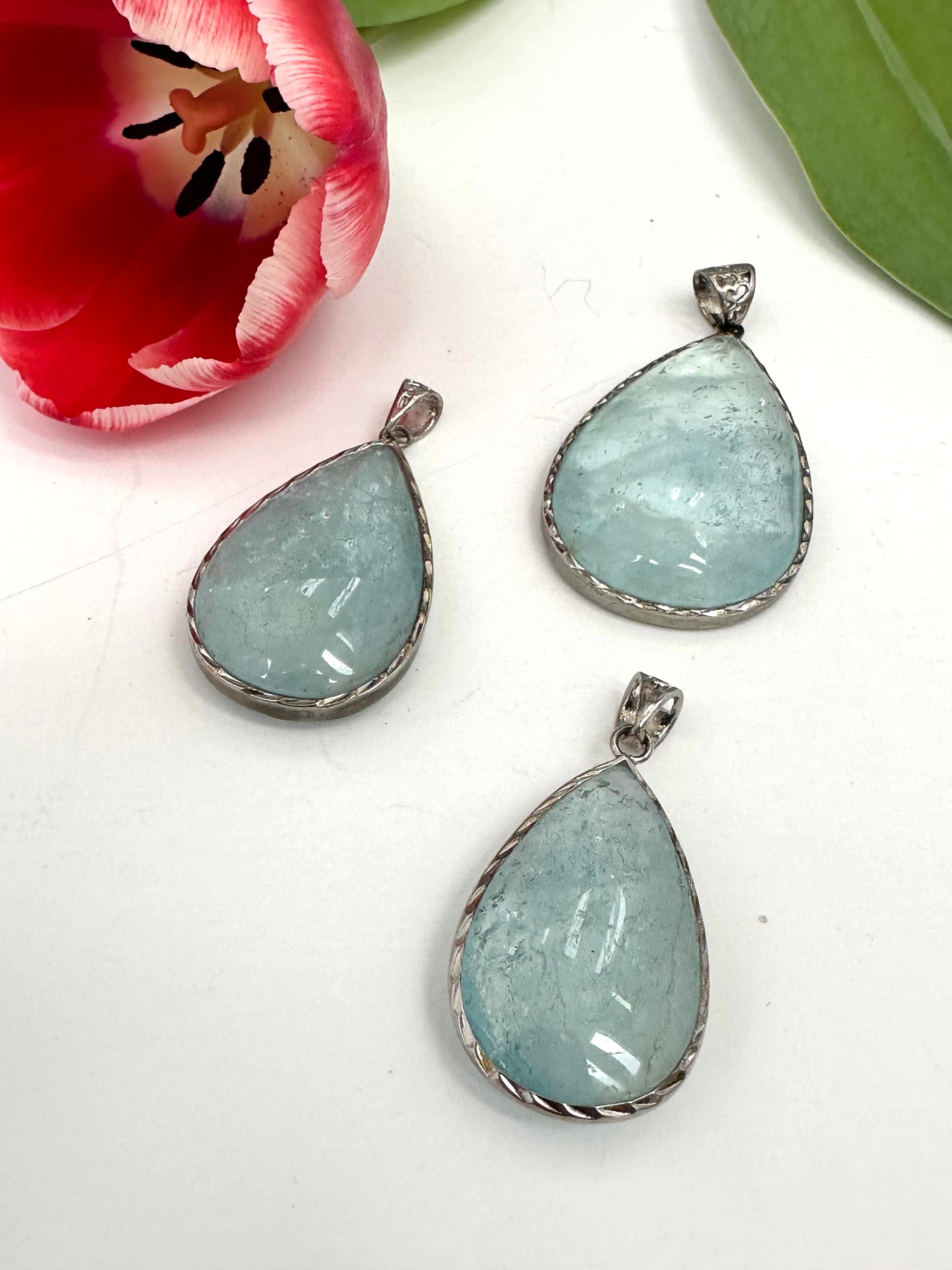 Three light blue teardrop-shaped pendants on a white surface with a pink flower in the background.