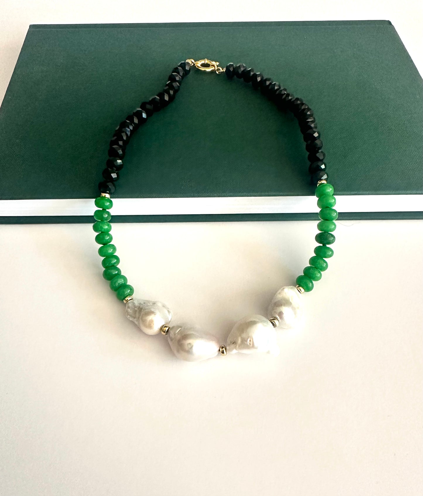 A beaded necklace with black faceted beads, green round beads, and 4 large white baroque pearls, displayed on a dark green rectangular box against a white background.