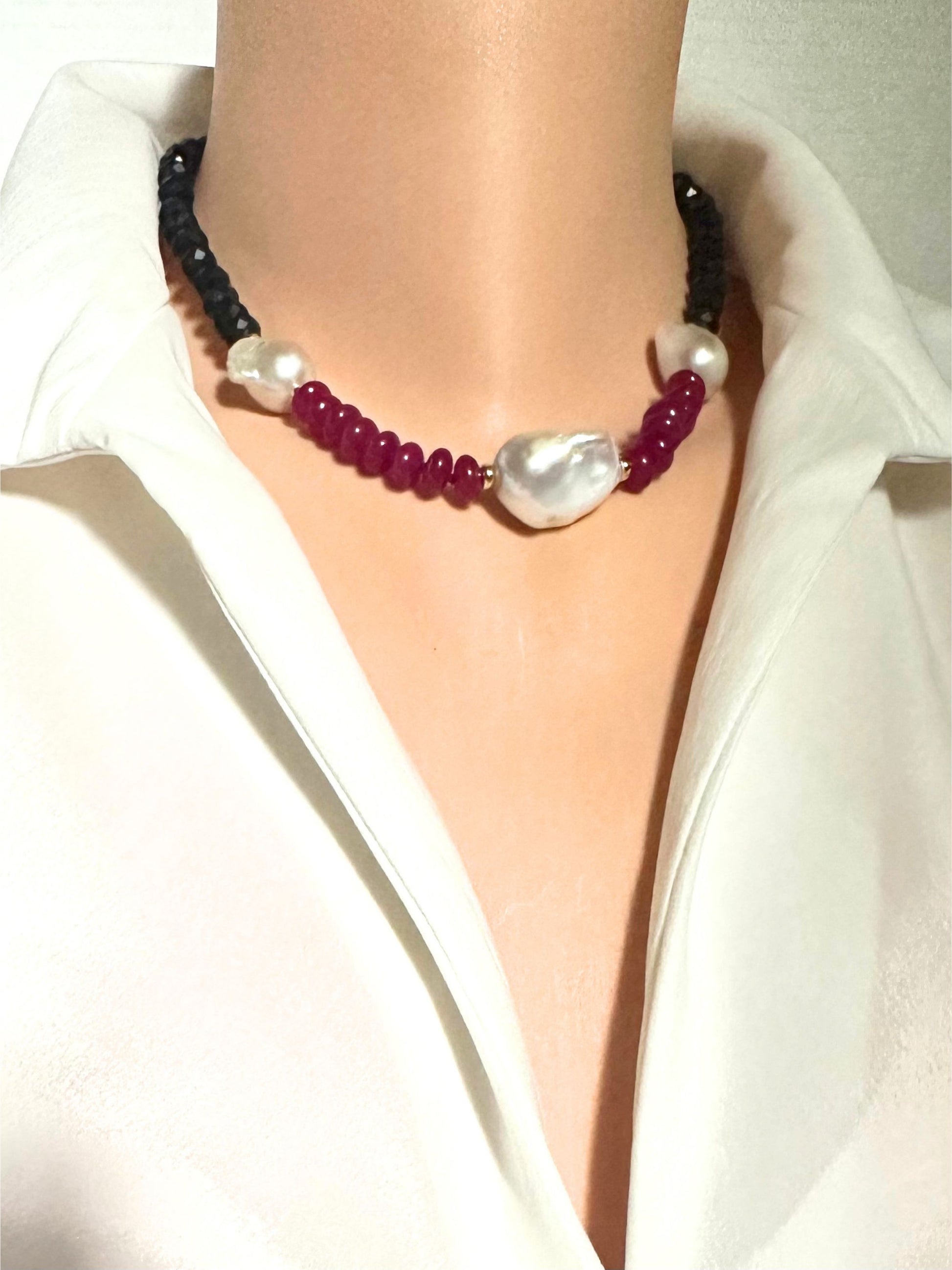 A close-up of a necklace worn on a mannequin torso dressed in a white collared shirt. The necklace features dark black beads, deep red beads, white pearl-like round beads, and a large irregular white baroque pearl at the center.