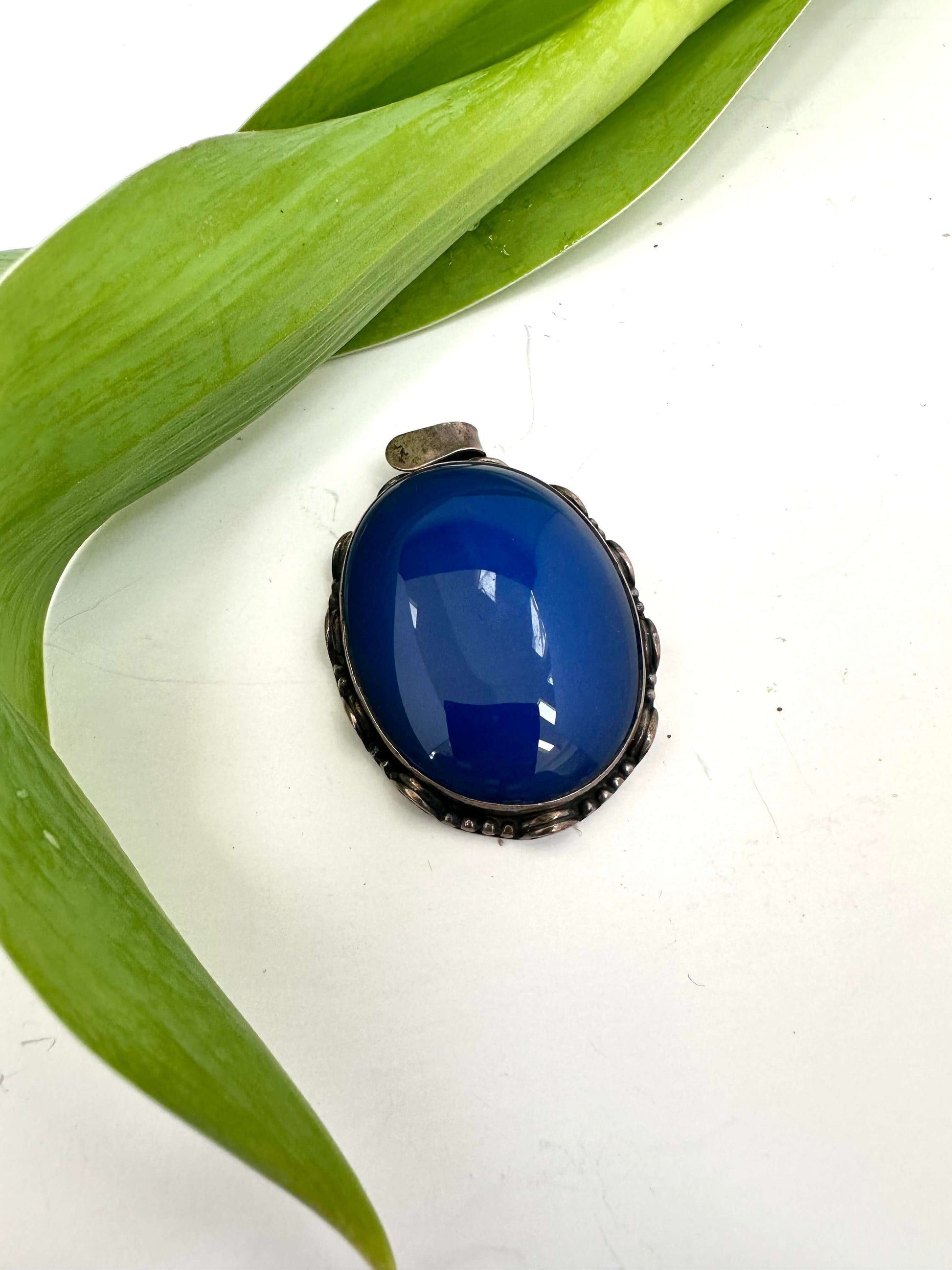 Vintage blue agate pendant in oxidized sterling silver with a deep cobalt-blue hue resting on a green leaf.