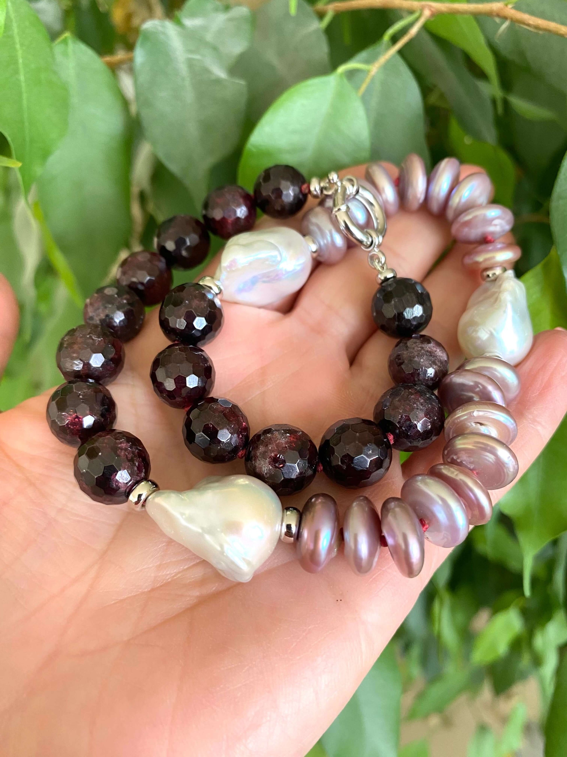Garnet pearl necklace featuring Baroque and coin pearls with rich gemstone colors, showcased in a hand against greenery.