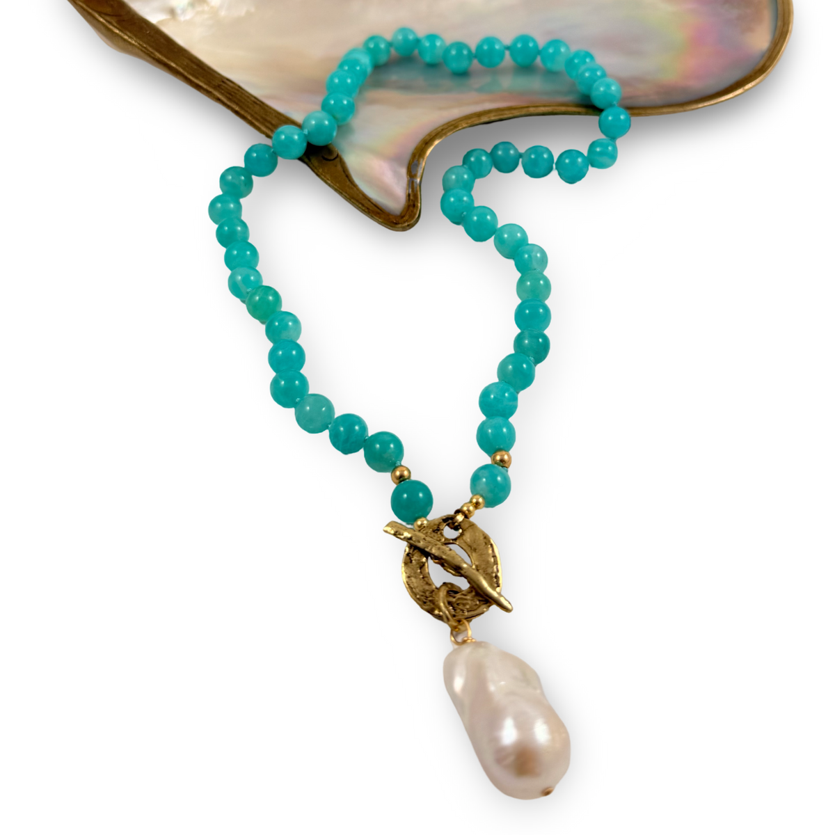 A beaded necklace with turquoise-blue round beads, gold-toned accents, and a large white baroque pearl pendant, displayed on a white background with a mother-of-pearl shell prop.