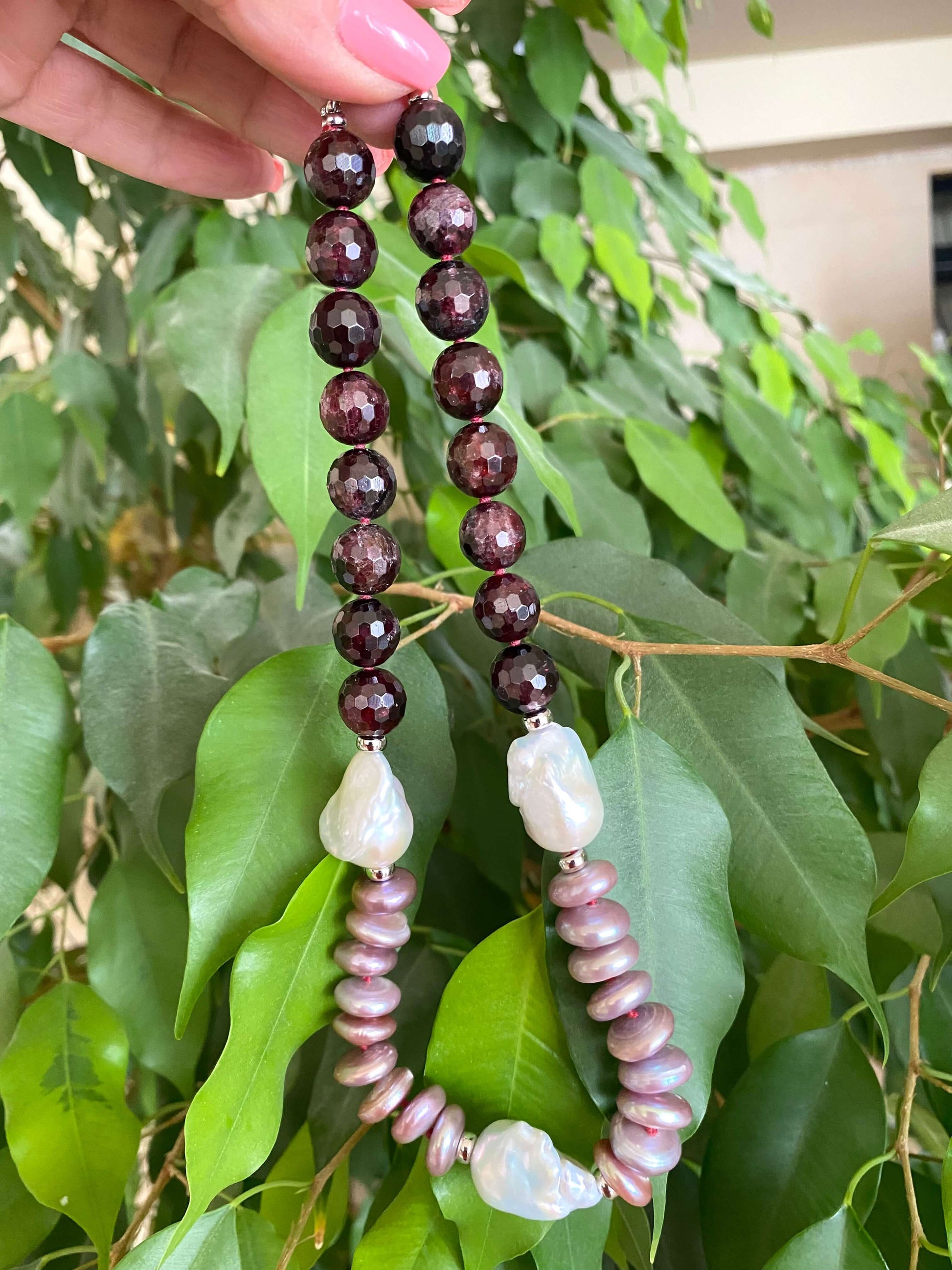 Garnet pearl necklace featuring faceted round garnet beads and Baroque and coin pearls, held against a leafy backdrop.