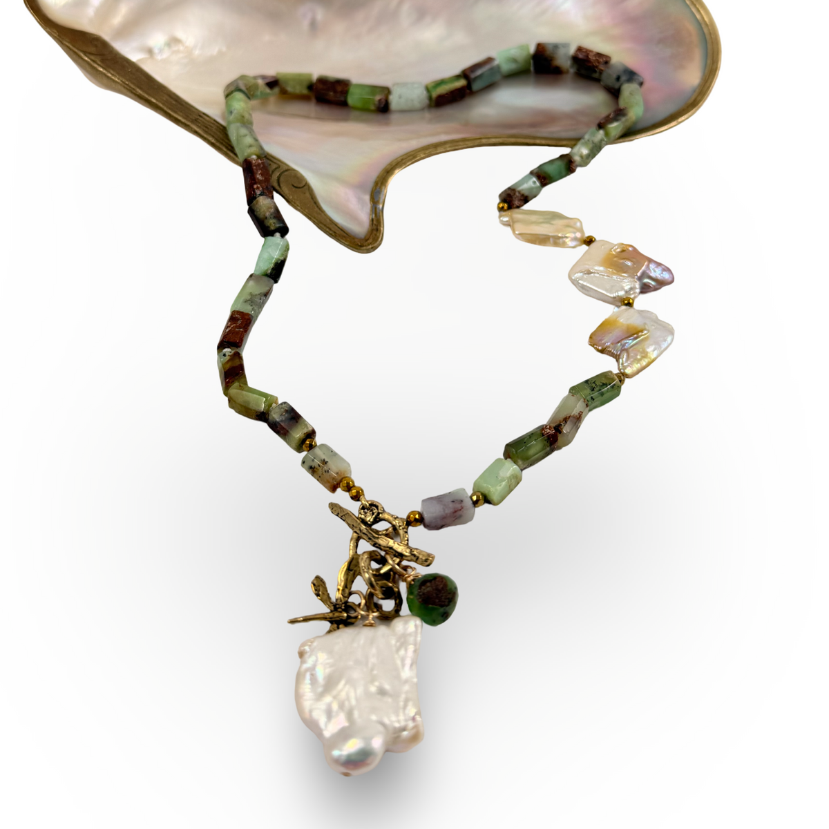 A handmade beaded necklace with a baroque pearl pendant, displayed on a mother-of-pearl clam shell. The necklace features green and brown beads, clear crystal chips, and a gold branch charm.