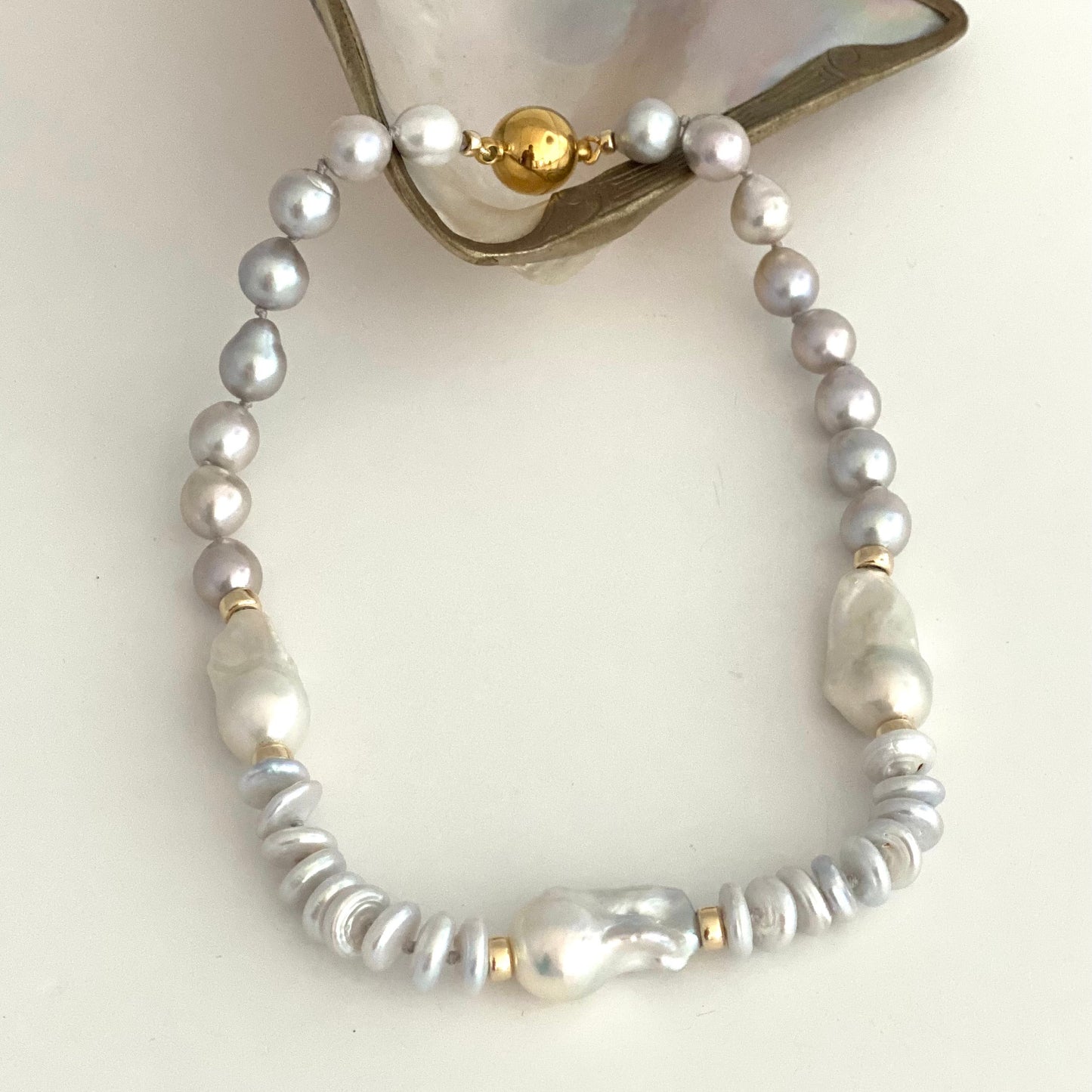 HAND KNOTTED NECKLACE made of white and silver baroque pearls, silver PEARL spacer beads, and gold bead, displayed on a white surface with a piece of mother-of-pearl shell at the top.