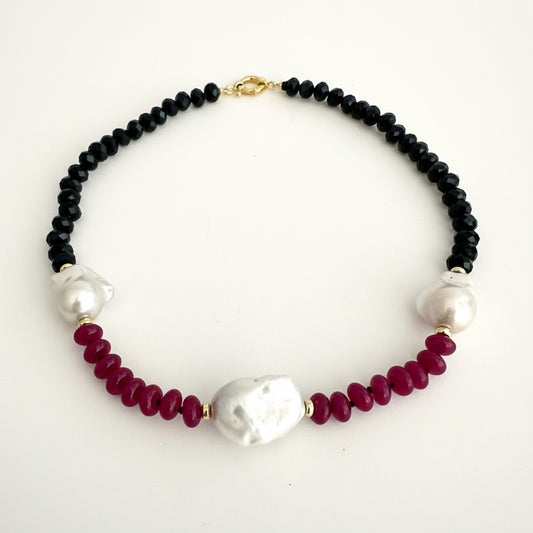 A beaded necklace featuring black faceted beads, deep red faceted beads, and three white baroque pearls, accented with small gold spacers and a gold clasp, shown on a plain white background.