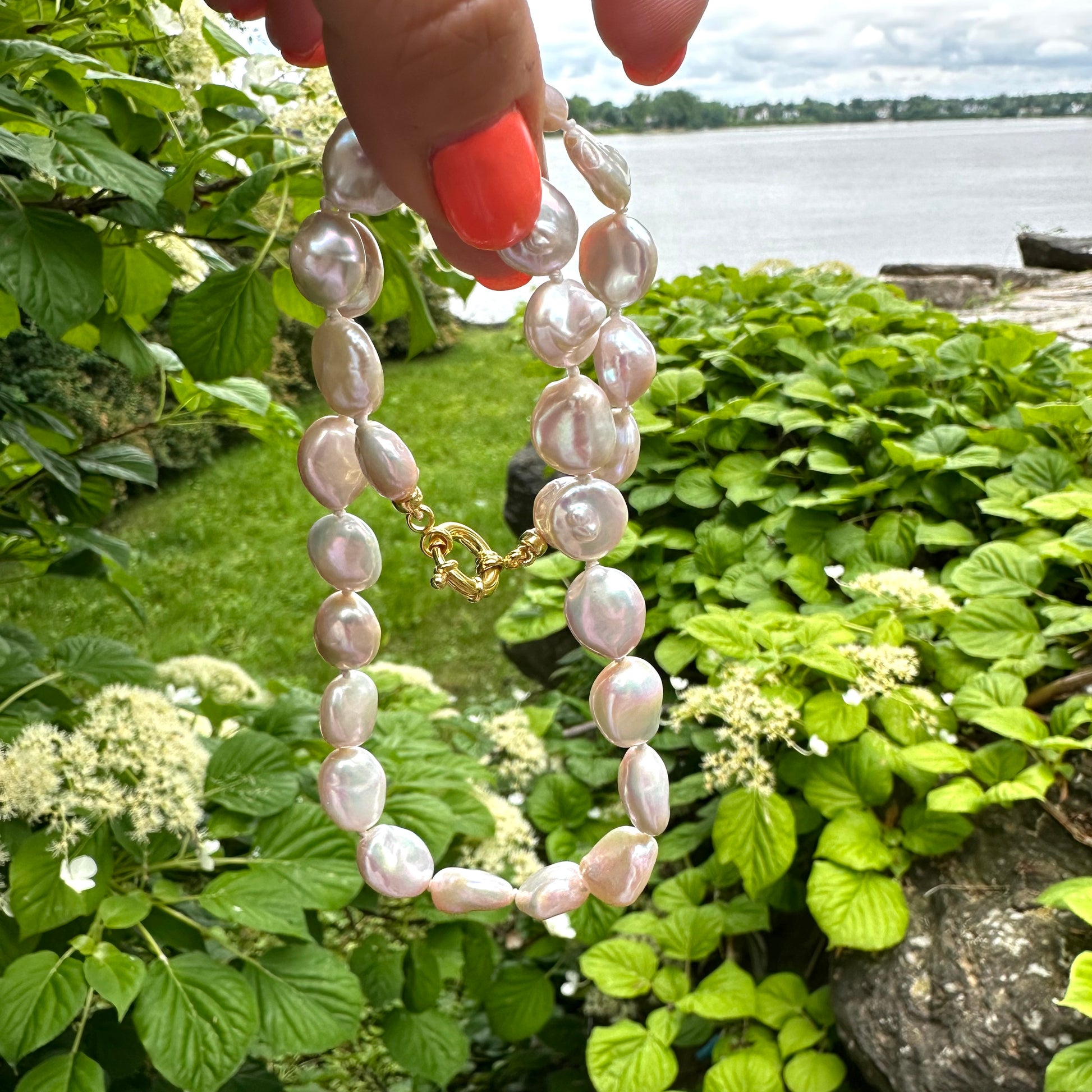 Freshwater coin pearl necklace held in hand with lake and greenery in background