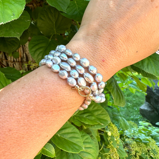 Grey nugget pearl necklace worn as bracelet on woman's wrist against green foliage