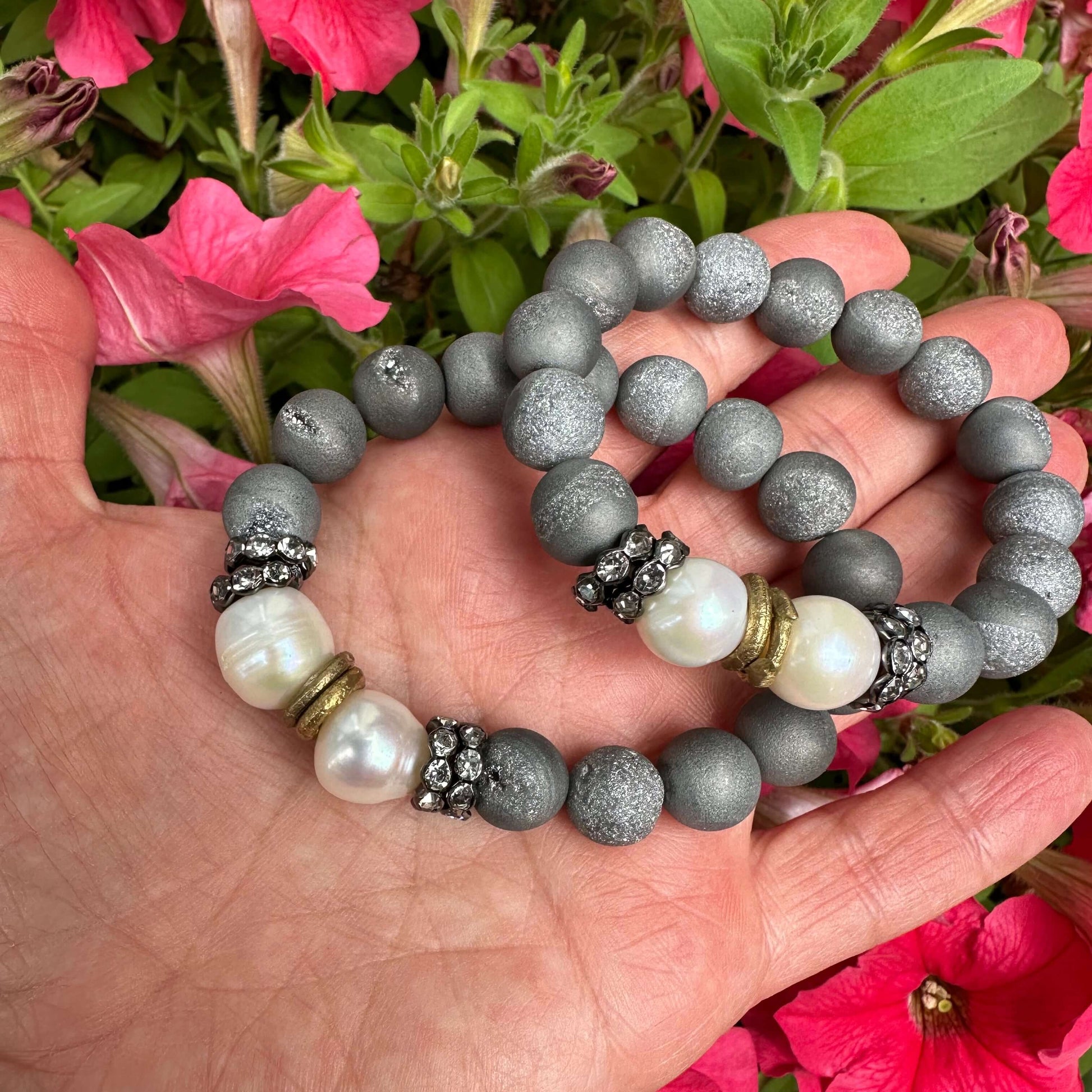 Two grey druzy agate and pearl bracelets held in hand against pink flowers