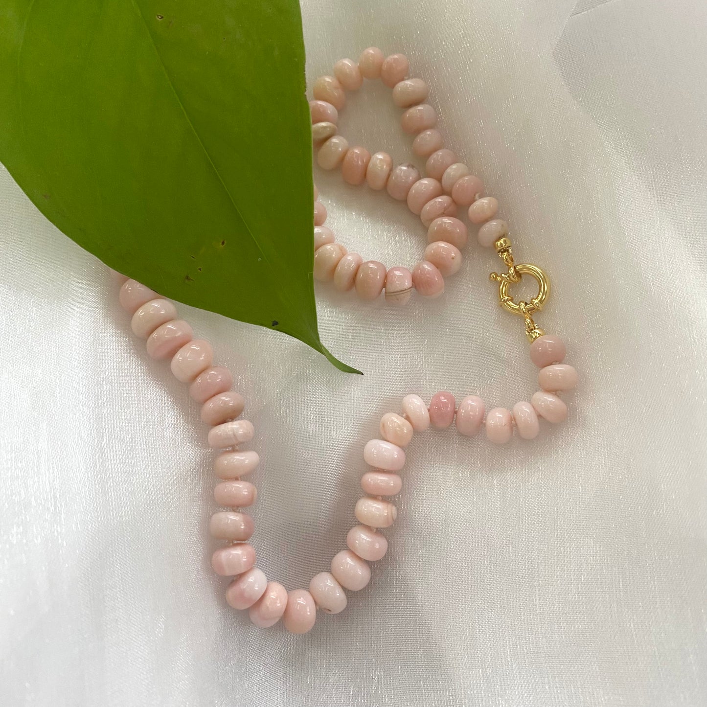 Graduated Pink Opal Necklace Gold flat lay on white fabric with green leaf, hand-knotted opal candy strand