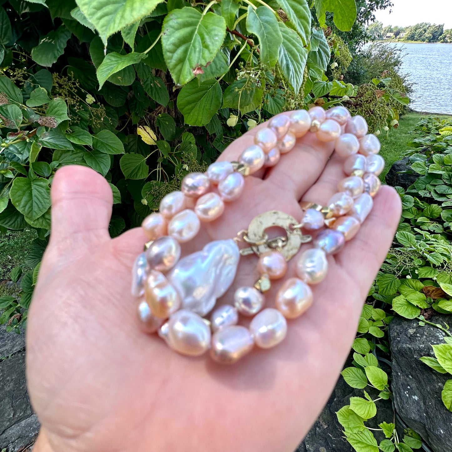 Pastel pearl necklace held in hand against green leaves and lake backdrop, close-up view