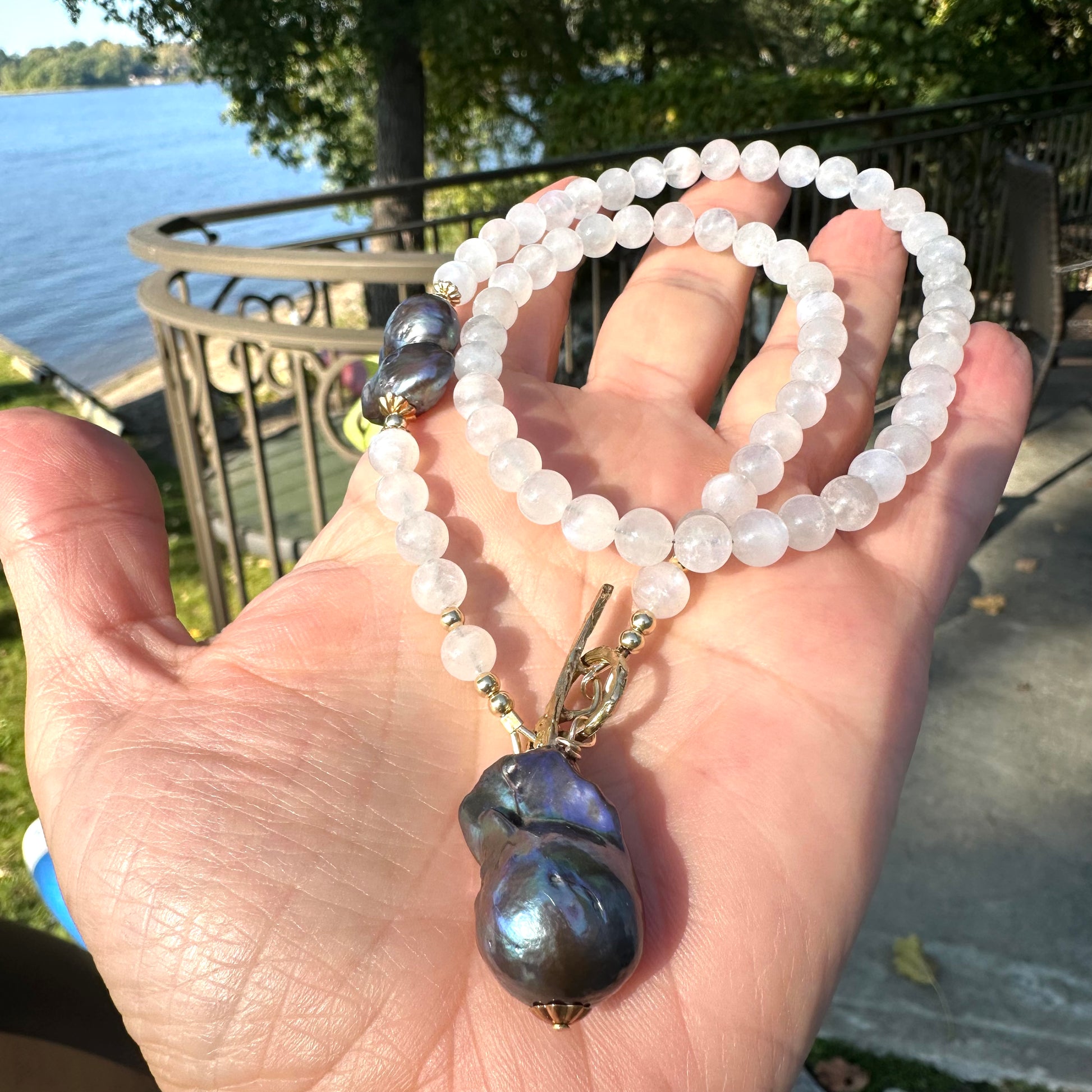 Moonstone necklace with black baroque pearl, handmade in Canada, displayed by a person near a scenic waterfront.