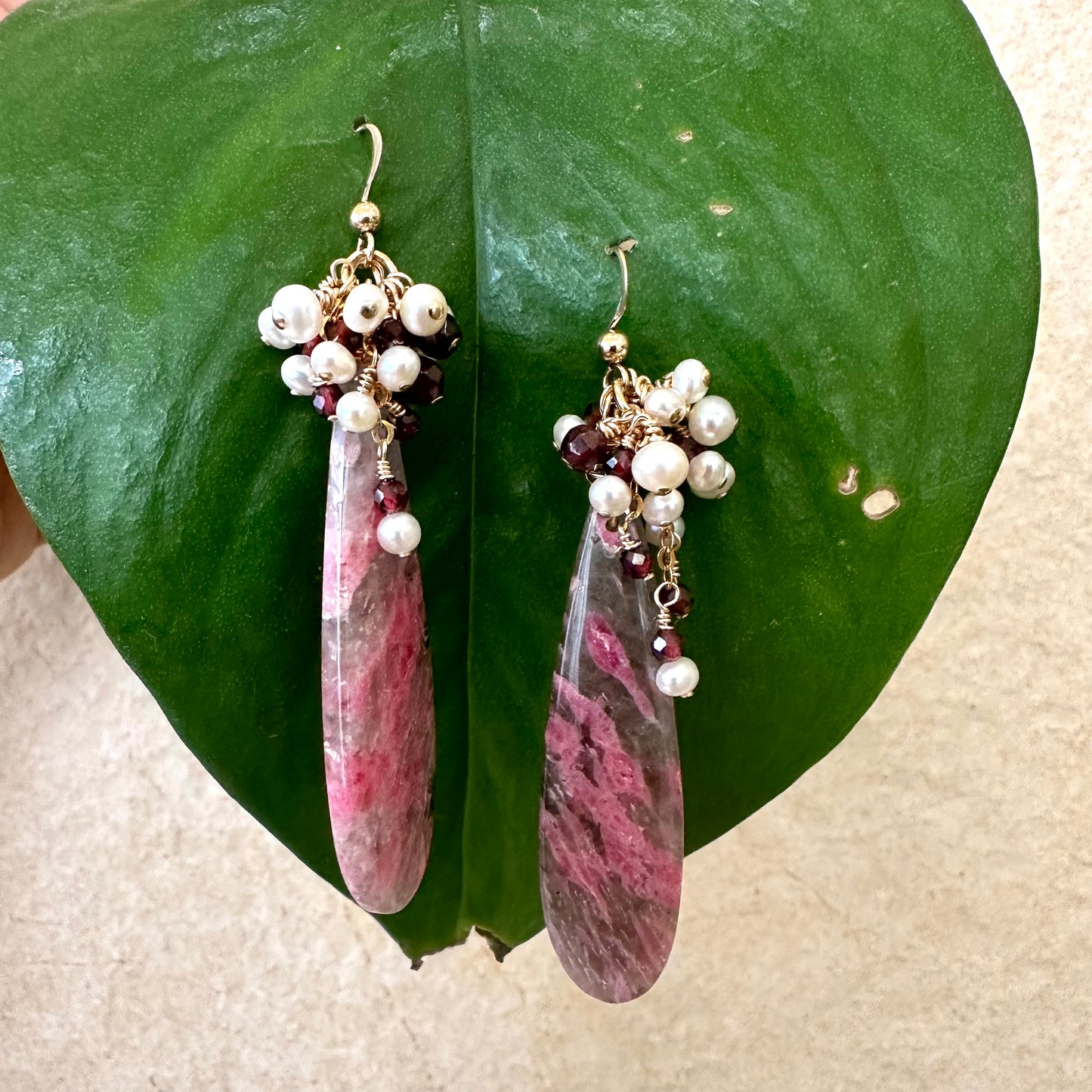 Pink rhodonite teardrop earrings with garnet and pearl cluster against green leaves handmade in Canada