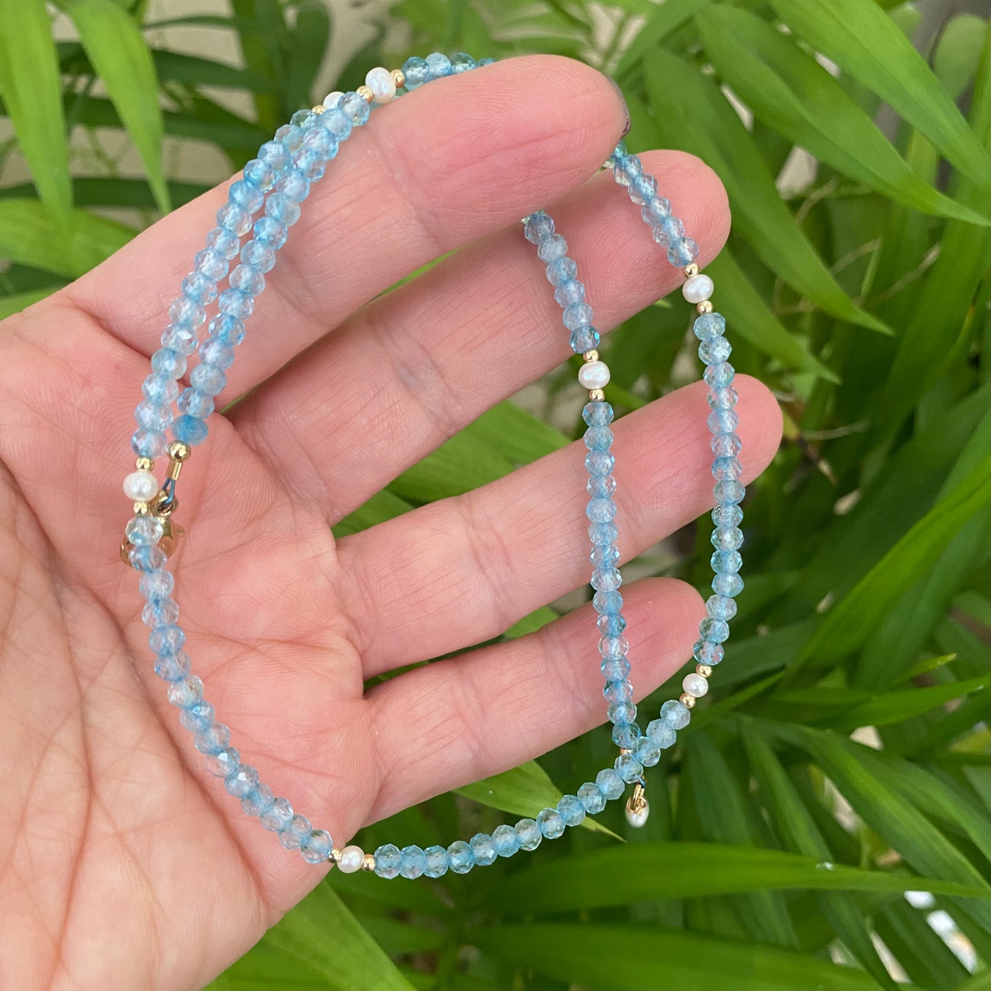 Blue topaz pearl choker necklace held in hand against green leaves