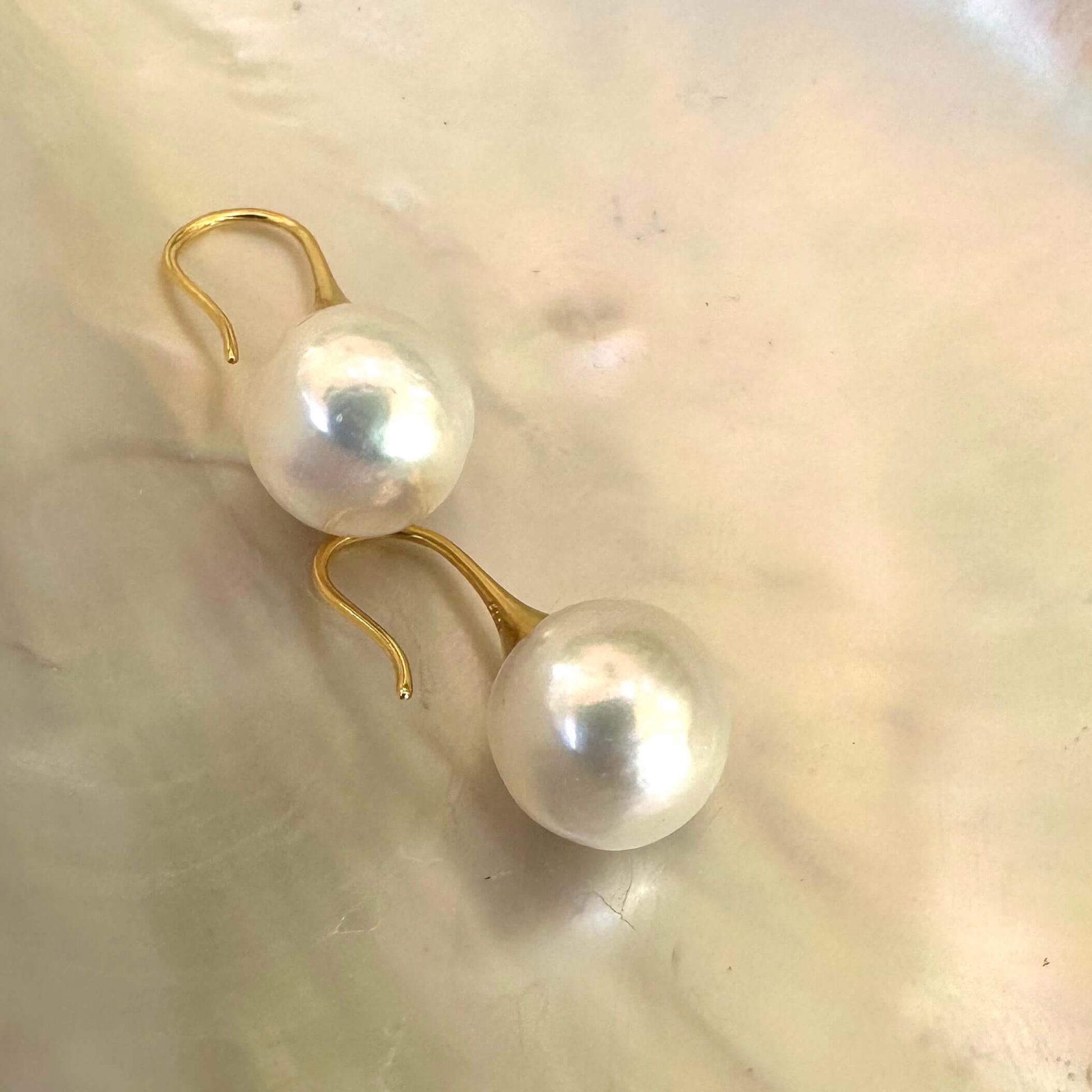 White pearl drop earrings on pearl shell background, showing near-round Edison pearls and gentle rippled surface texture.