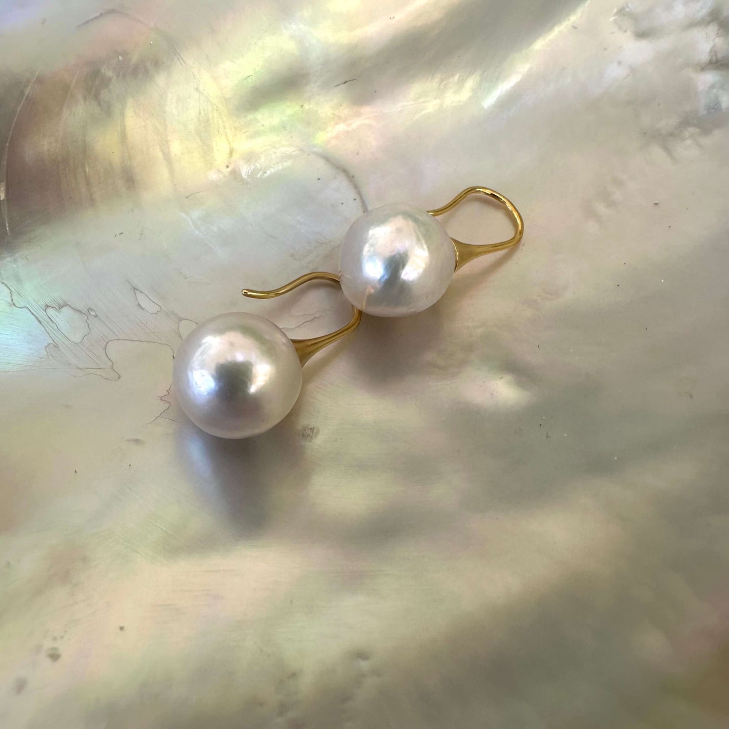 White pearl drop earrings with large round Edison pearls resting on shell, showing mirror-like luster and gold finish.