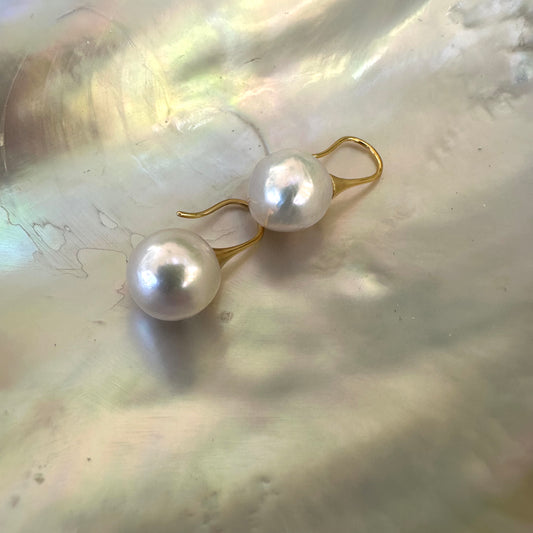 White pearl drop earrings with large round Edison pearls resting on shell, showing mirror-like luster and gold finish.