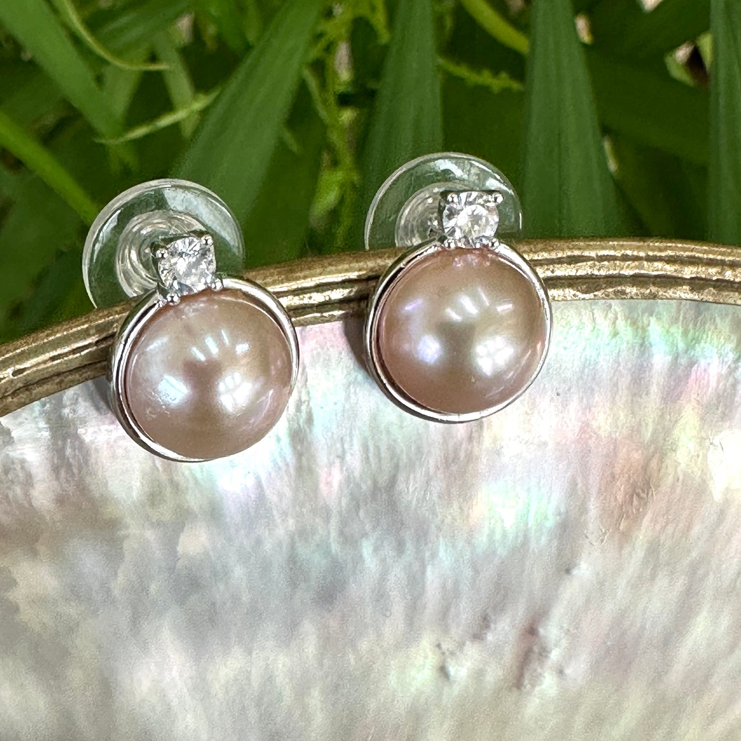 Pink mabe pearl stud earrings with CZ in sterling silver, close-up dangling from white pearl shell with green foliage background.