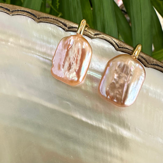 Golden peach pearl drop earrings on shell among foliage, highlighting radiant peach color and artistic baroque pearl detail.
