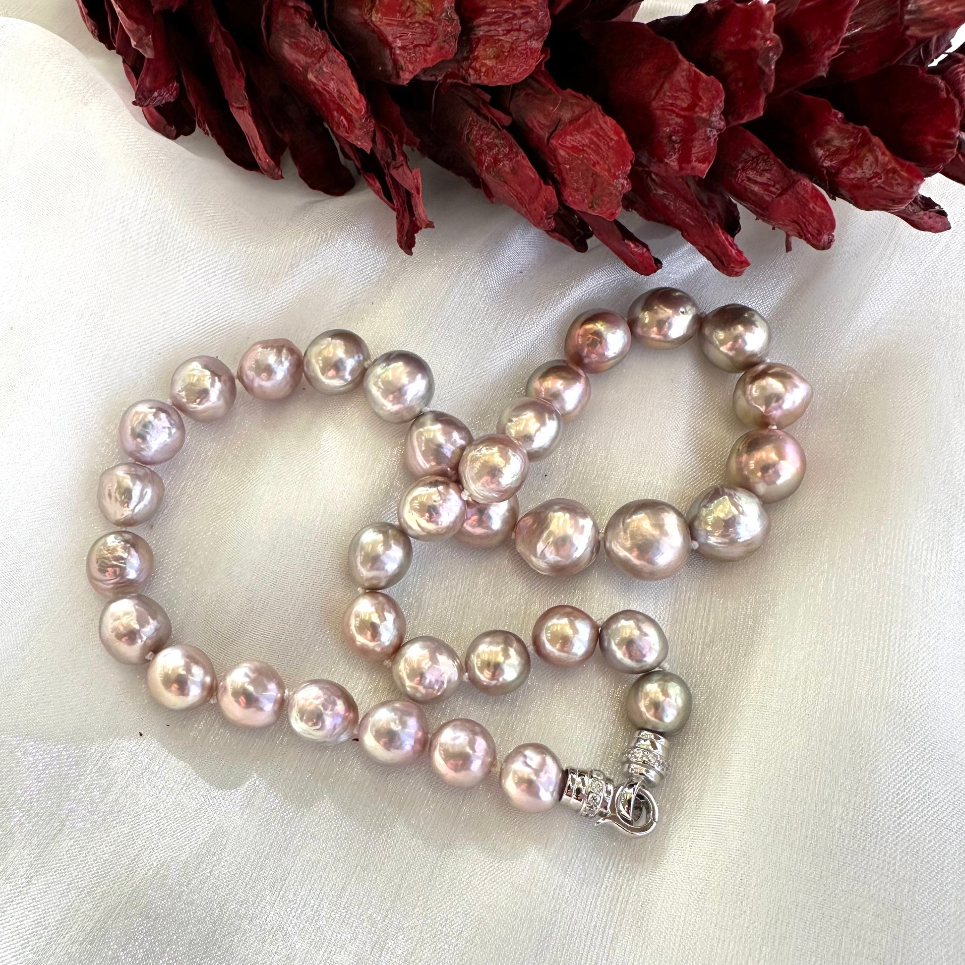 Pink lavender pearl necklace on white fabric beside red pine cone ornament, capturing holiday-ready warmth and shine.