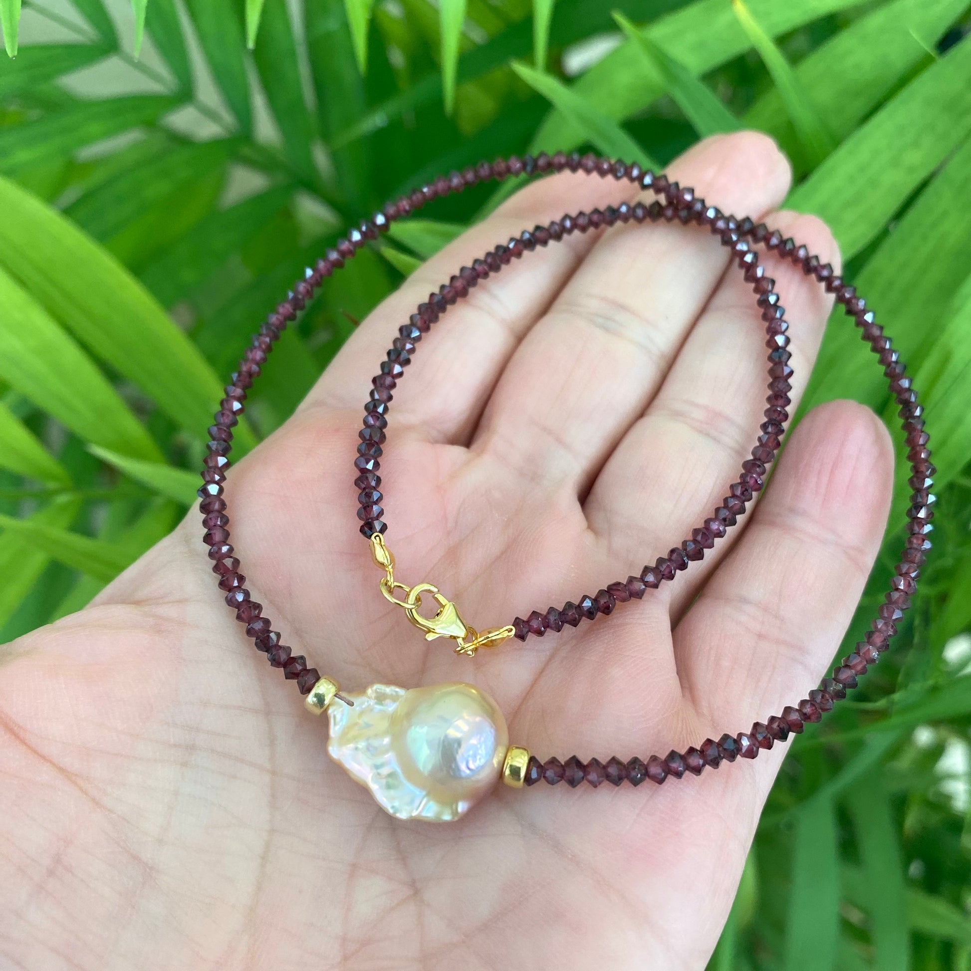 Red garnet baroque pearl necklace featuring golden pink pearl pendant, displayed in hand against lush green background.