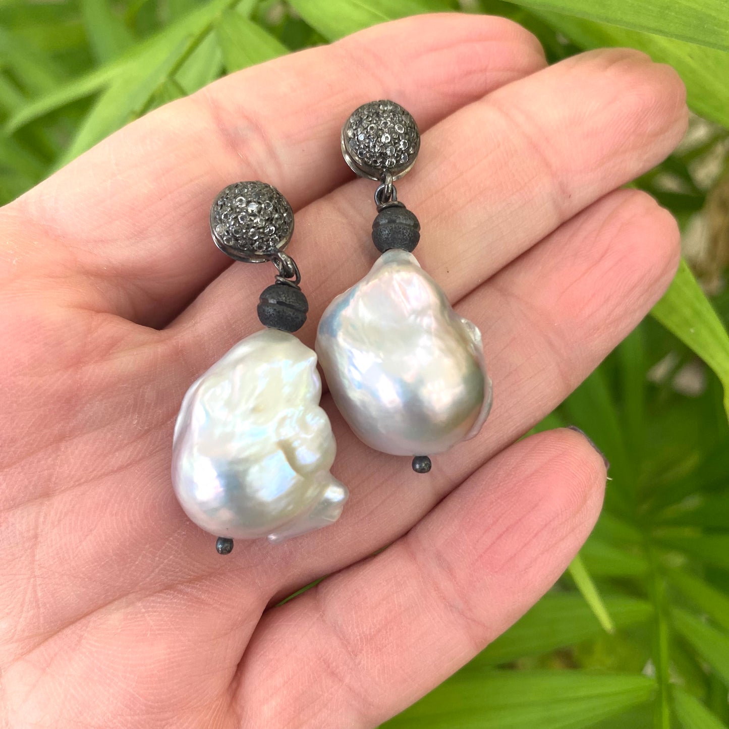 Baroque pearl drop earrings with diamond pavé studs in oxidized silver, showcased in a hand.