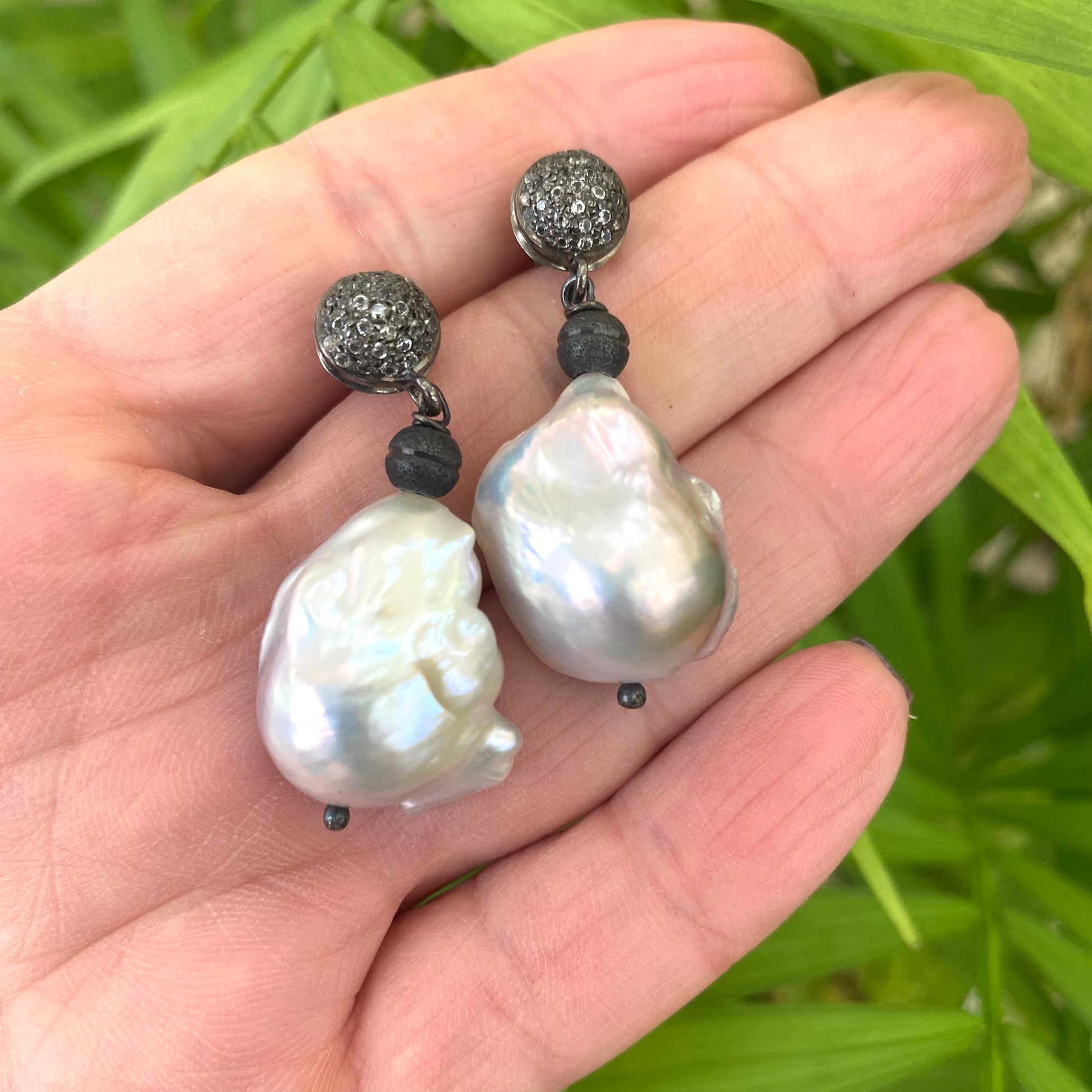 Baroque pearl drop earrings with diamond pavé studs in oxidized silver, showcased in a hand.