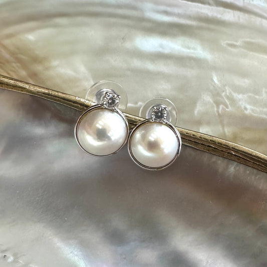White mabe pearl stud earrings hanging from white shell, showing round Edison pearls with mirror-like sheen and CZ sparkle.