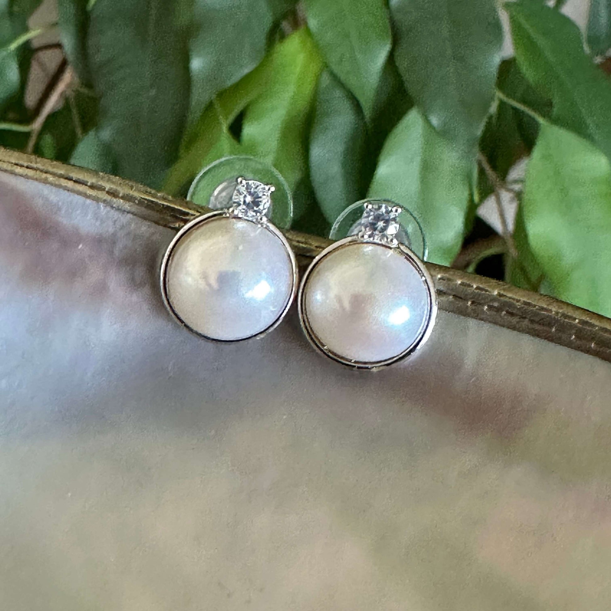 White mabe pearl stud earrings with CZ on white shell and green leaves, reflecting natural light and silver-white radiance.