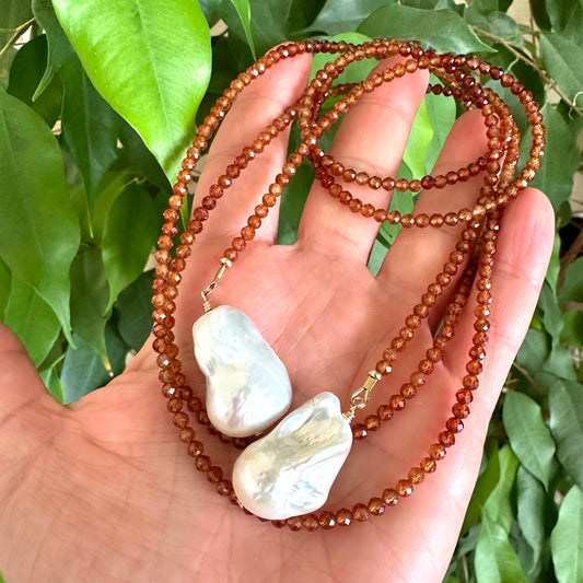 Hessonite Garnet and Baroque Pearl Lariat Necklace held in hand against green leaf backdrop, handcrafted in Canada.