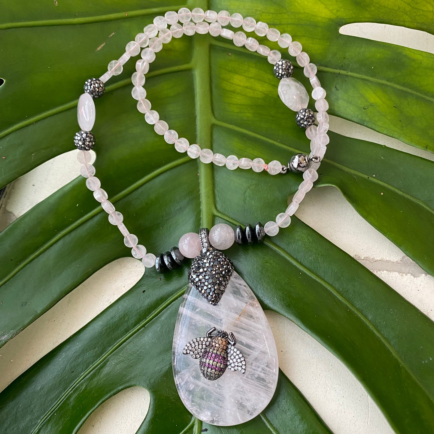 Rose quartz necklace featuring rhinestone pavé bee pendant on green leaf background.