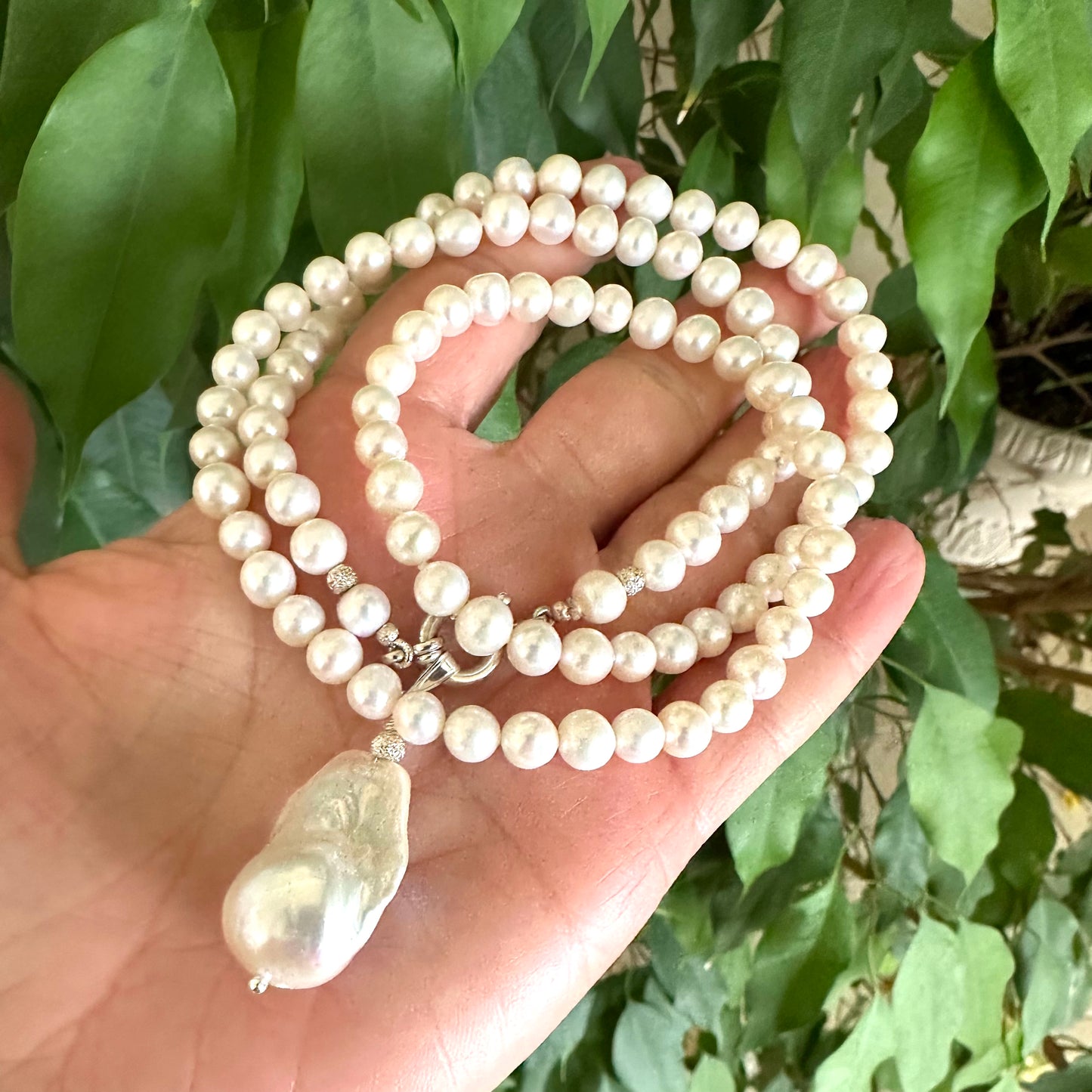 Hand holding white pearl necklace with baroque pendant near green leaf background