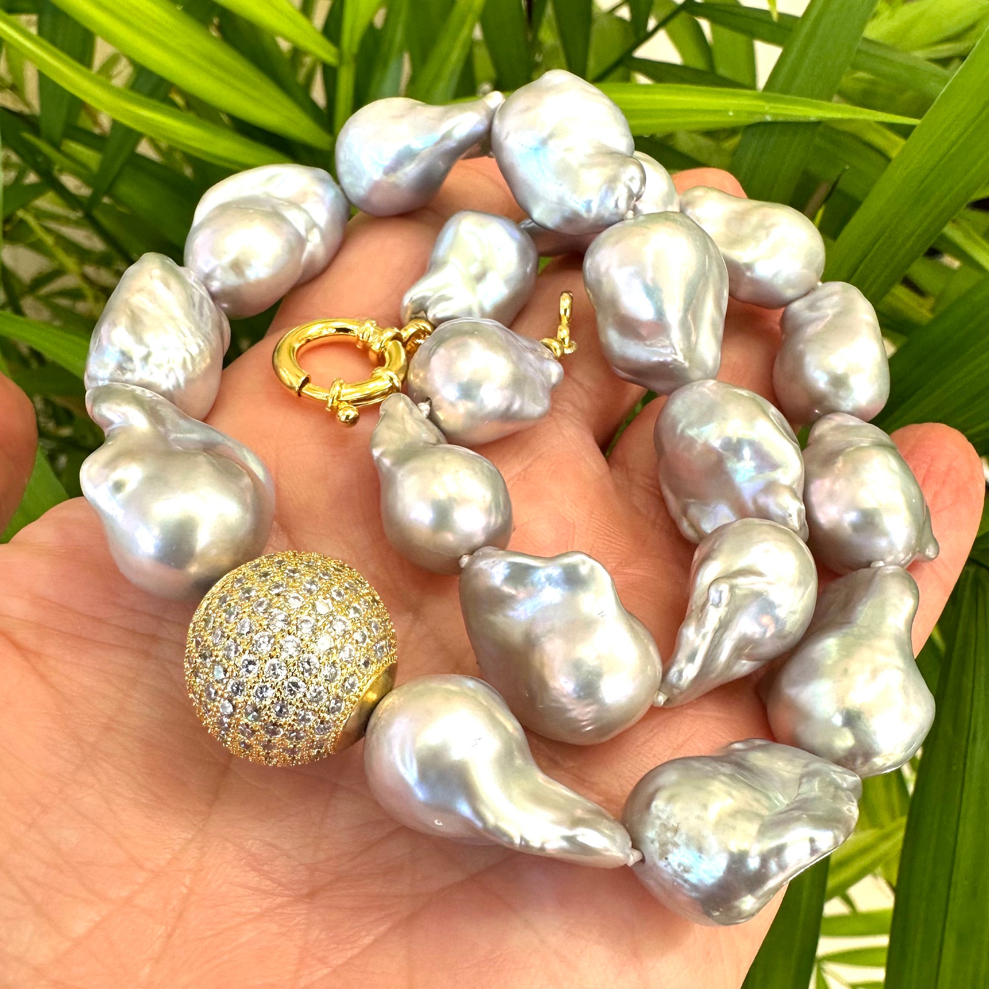 Silver grey baroque pearl necklace held in hand against green leaf background