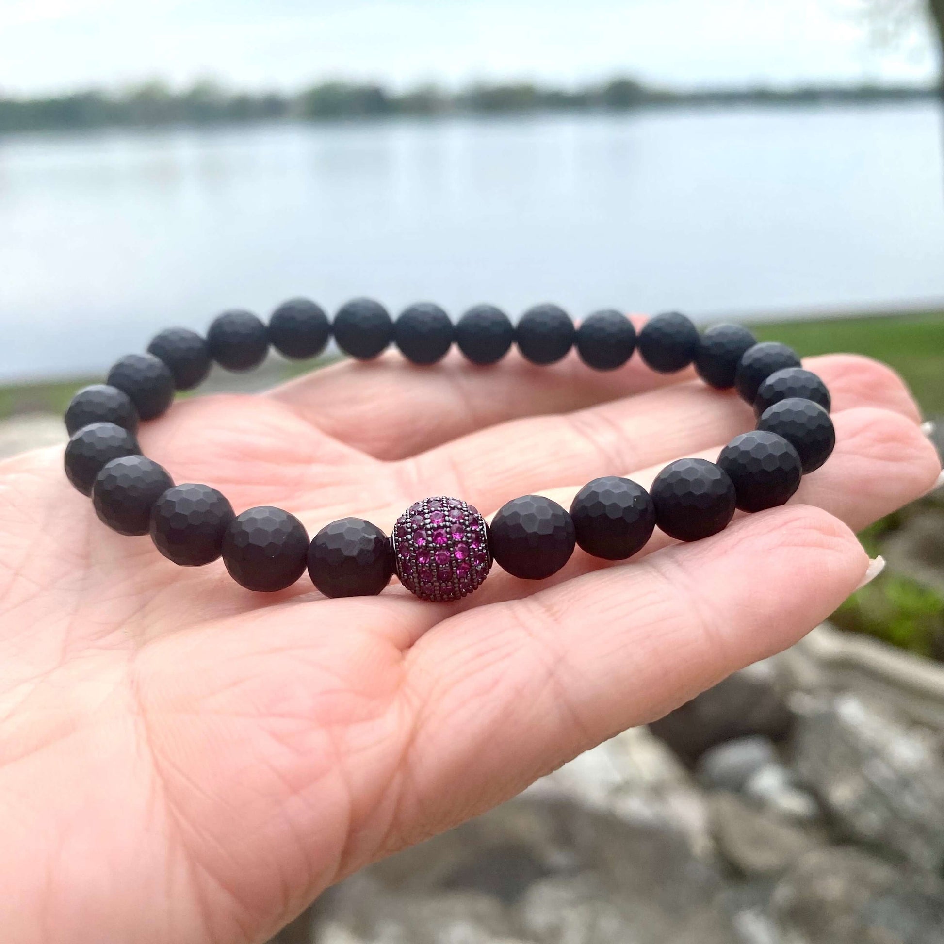 Matte black onyx mens bracelet featuring a ruby red CZ center spacer, displayed on hand by the water.