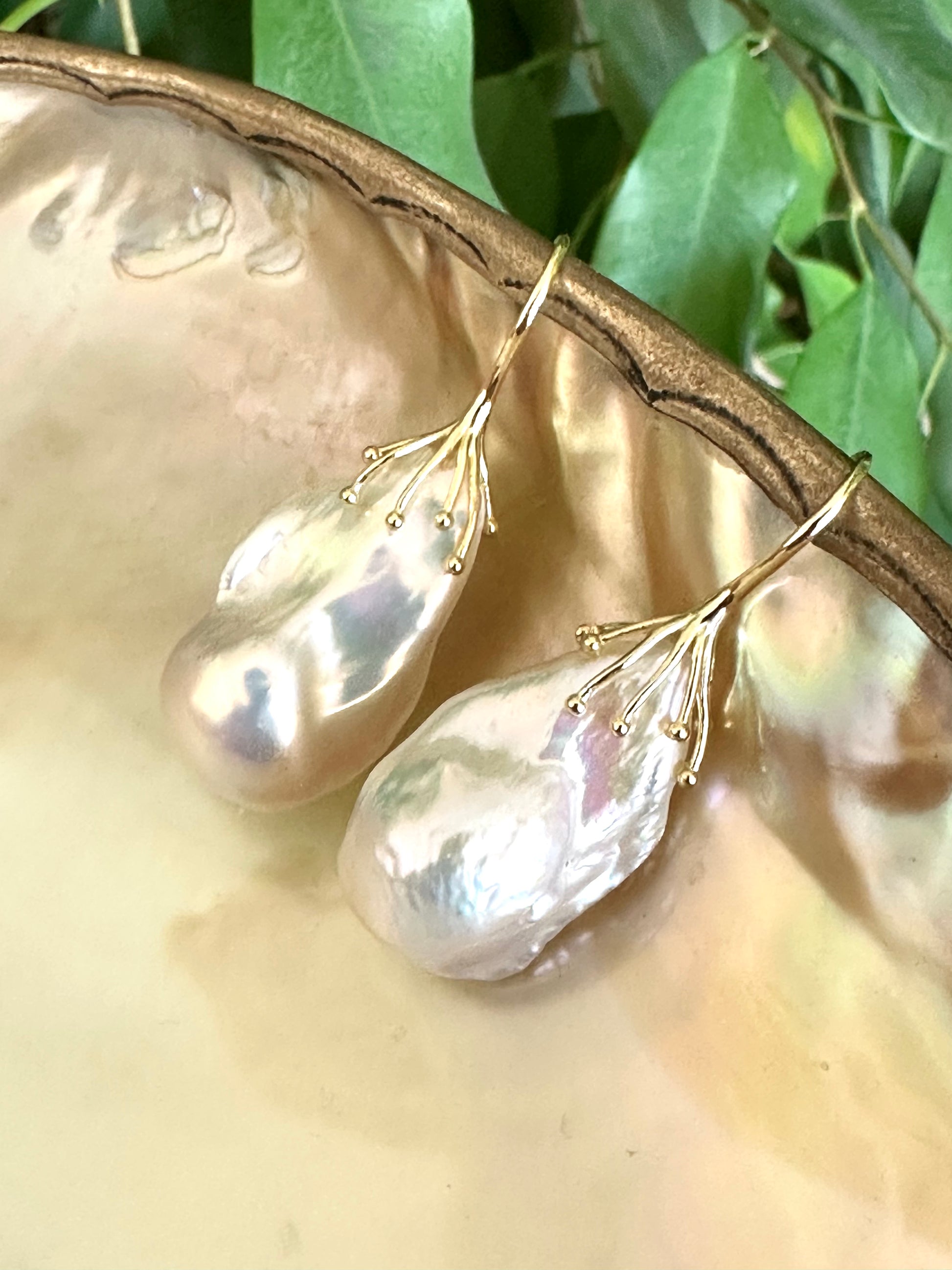 White baroque pearl drop earrings resting on green leaf and pearl shell backdrop, silver hooks visible