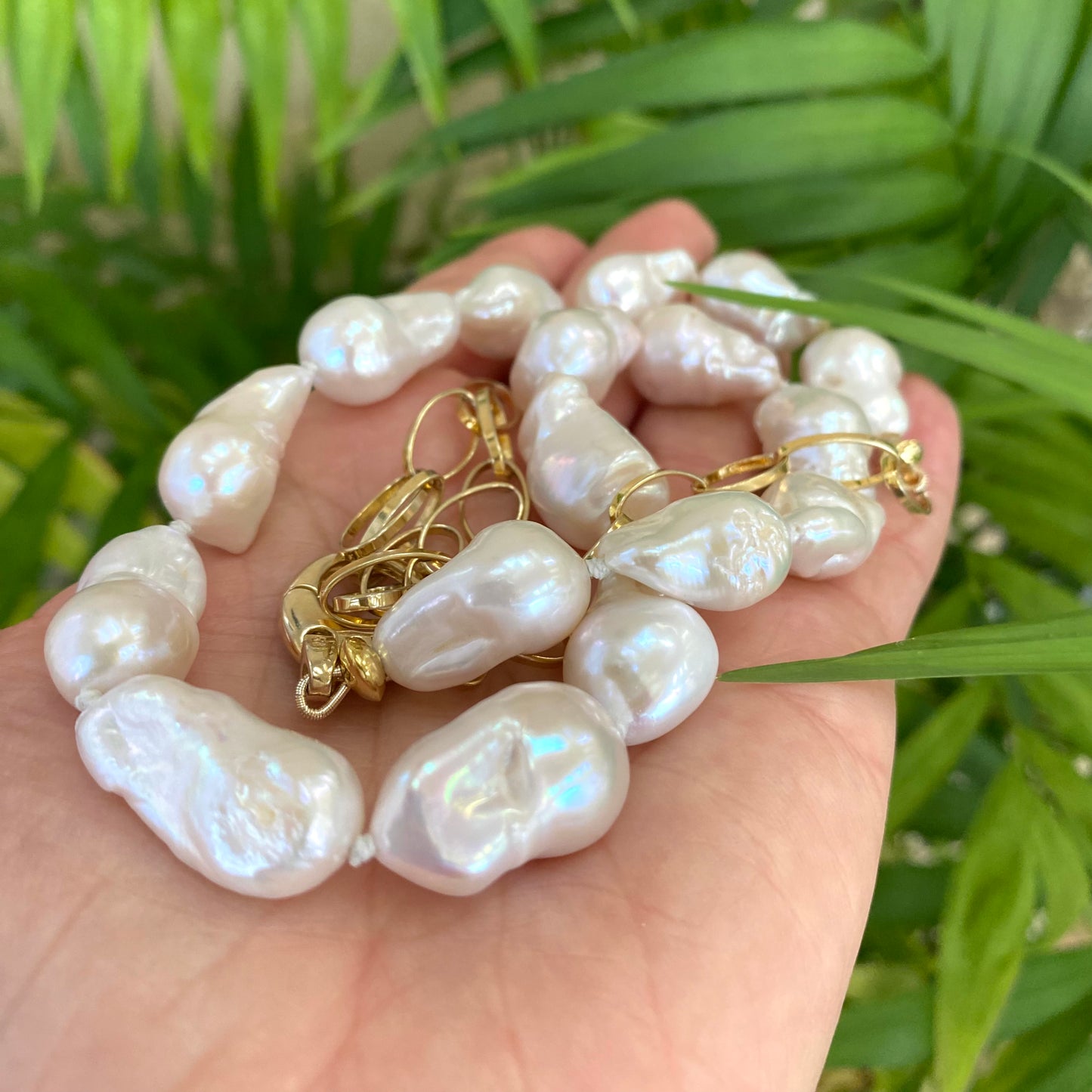 White baroque pearl and gold chain necklace held in hand against green foliage, designed and handmade in Canada.