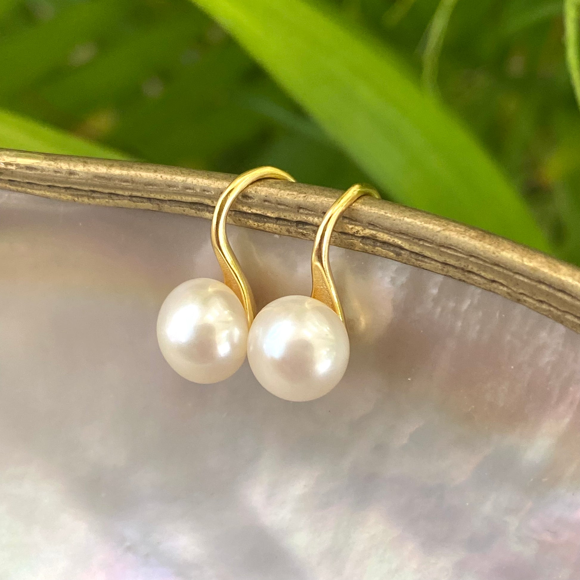 Button Pearl Earrings – gold pair displayed on pearl shell with green leaf backdrop
